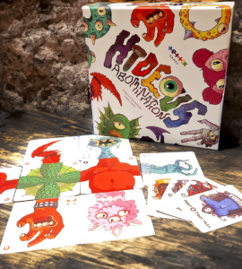 Colorful board game "HIDEOUS Abominations" displayed on a rustic wooden table, featuring unique character cards and a creatively designed box, set against a textured stone backdrop.