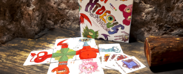 Colorful board game "HIDEOUS Abominations" displayed on a rustic wooden table, featuring unique character cards and a creatively designed box, set against a textured stone backdrop.