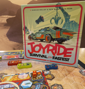 Board game "Joyride: Survival of the Fastest" featuring vibrant artwork of a futuristic car and game components displayed on a tabletop with a desert background. The game includes colorful vehicle tokens and strategic elements for players to engage in high-speed challenges.