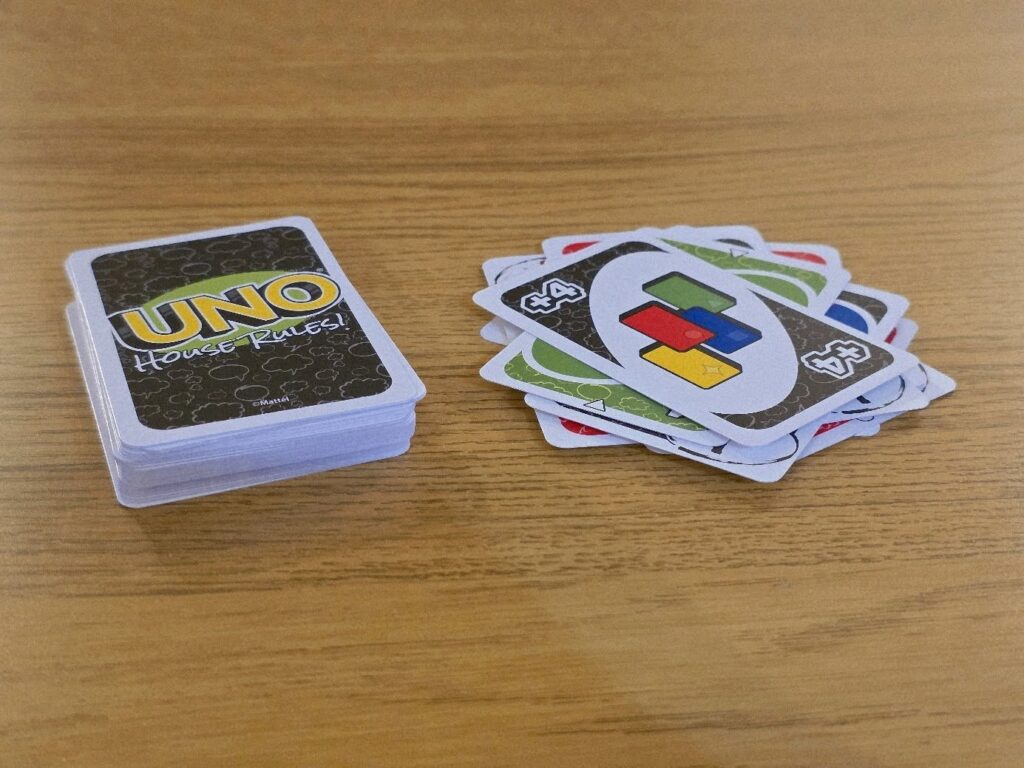 UNO card game featuring a stack of house rules cards next to a pile of colorful playing cards, including a "+4" card, displayed on a wooden table. Ideal for family game nights and gatherings.