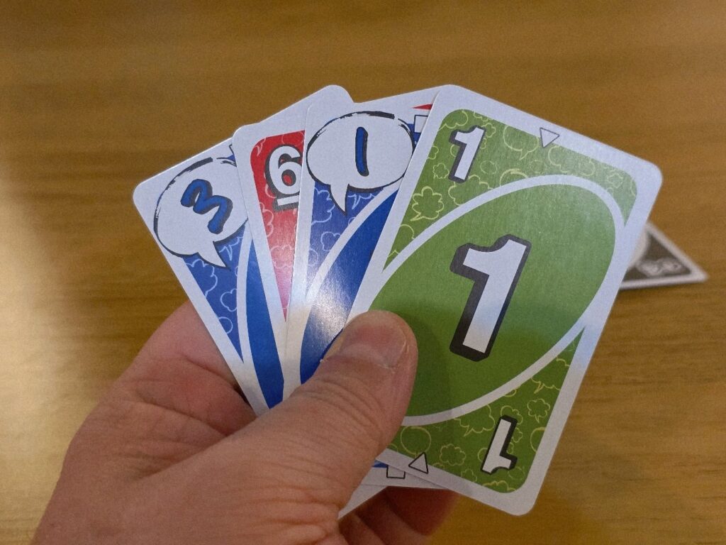 Image of a hand holding four colorful playing cards, displaying the numbers 1, 3, and 6, with distinct color backgrounds of blue, red, and green. The cards are laid out on a wooden table, showcasing a casual gaming scenario.