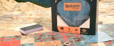 Trailblazers Travel Edition game box displayed on a rocky surface, surrounded by game cards featuring outdoor illustrations, suitable for 1-4 players and ages 8 and up. Ideal for road trips and family fun, emphasizing engagement with nature.