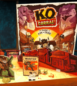 Colorful board game "K.O. Corral" displayed on a wooden table, featuring characters and a "Wanted" poster for Claude VonSnap, set in a Wild West theme with various game pieces and dice.