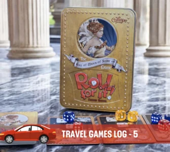 Alt text: A close-up of the board game "Roll for It!" Deluxe Edition displayed on a marble surface, featuring its distinctive gold tin box and colorful dice. Accompanying cards are spread out, showcasing artwork and game components, in a stylish setting with classical pillars in the background.