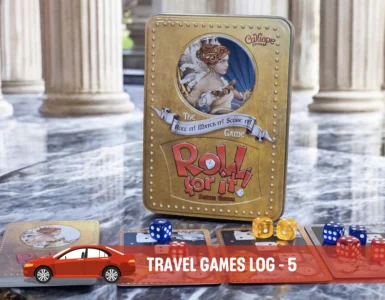 Alt text: A close-up of the board game "Roll for It!" Deluxe Edition displayed on a marble surface, featuring its distinctive gold tin box and colorful dice. Accompanying cards are spread out, showcasing artwork and game components, in a stylish setting with classical pillars in the background.