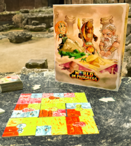 Board game "World in Progress" displayed on a stone table in an ancient outdoor setting, featuring colorful game tiles and a box with illustrated characters.