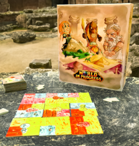 Board game "World in Progress" displayed on a stone table in an ancient outdoor setting, featuring colorful game tiles and a box with illustrated characters.