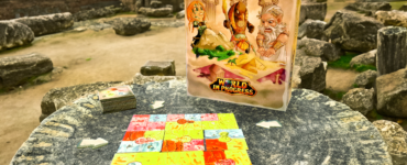 Board game "World in Progress" displayed on a stone table in an ancient outdoor setting, featuring colorful game tiles and a box with illustrated characters.