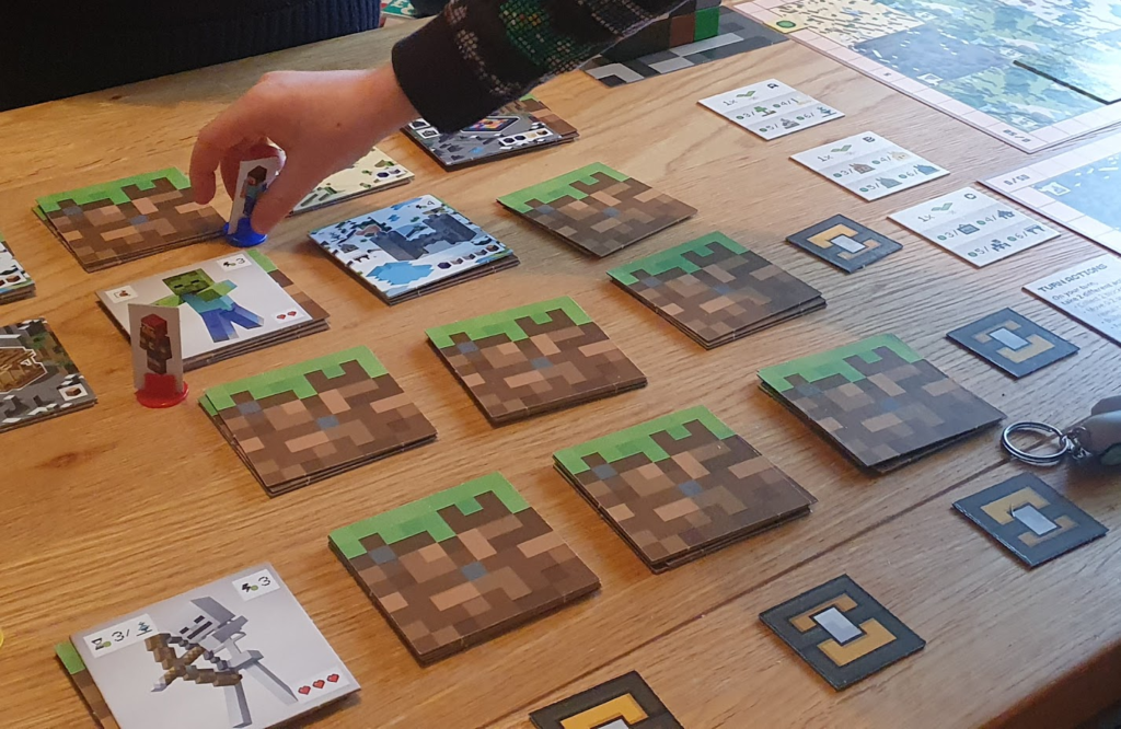 Image of a Minecraft-themed board game setup, featuring character cards and game pieces on a wooden table. A player's hand is seen placing a character piece near grassy block representations. Game cards with graphics of various characters, including a zombie and a skeleton, are positioned across the table, indicating active gameplay and strategy.