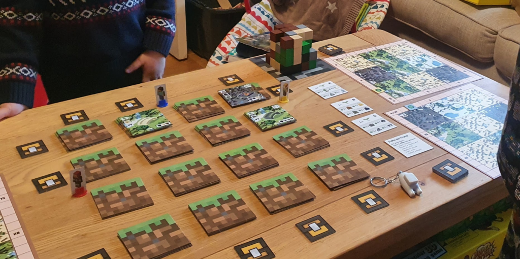 Tabletop gaming scene featuring a Minecraft-themed board game setup. The game includes colorful tiles resembling dirt blocks, player pieces, and reference cards laid out on a wooden table. A cozy living room background adds to the relaxed gaming atmosphere.