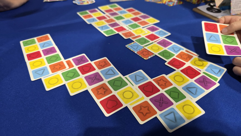Colorful game cards are arranged in a playful layout on a blue tablecloth, showcasing different shapes and symbols like hearts, stars, and geometric figures. A hand is seen holding a card, indicating a strategic move in the game. The vibrant design highlights the engaging nature of tabletop gaming.