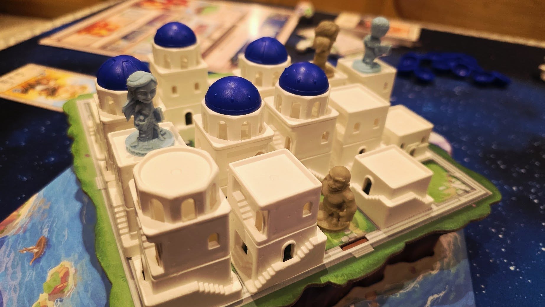 Colorful game board featuring a three-dimensional model of a Greek island with white buildings and blue domes, along with various game pieces positioned strategically. Perfect for family game night or enthusiasts of strategy board games.