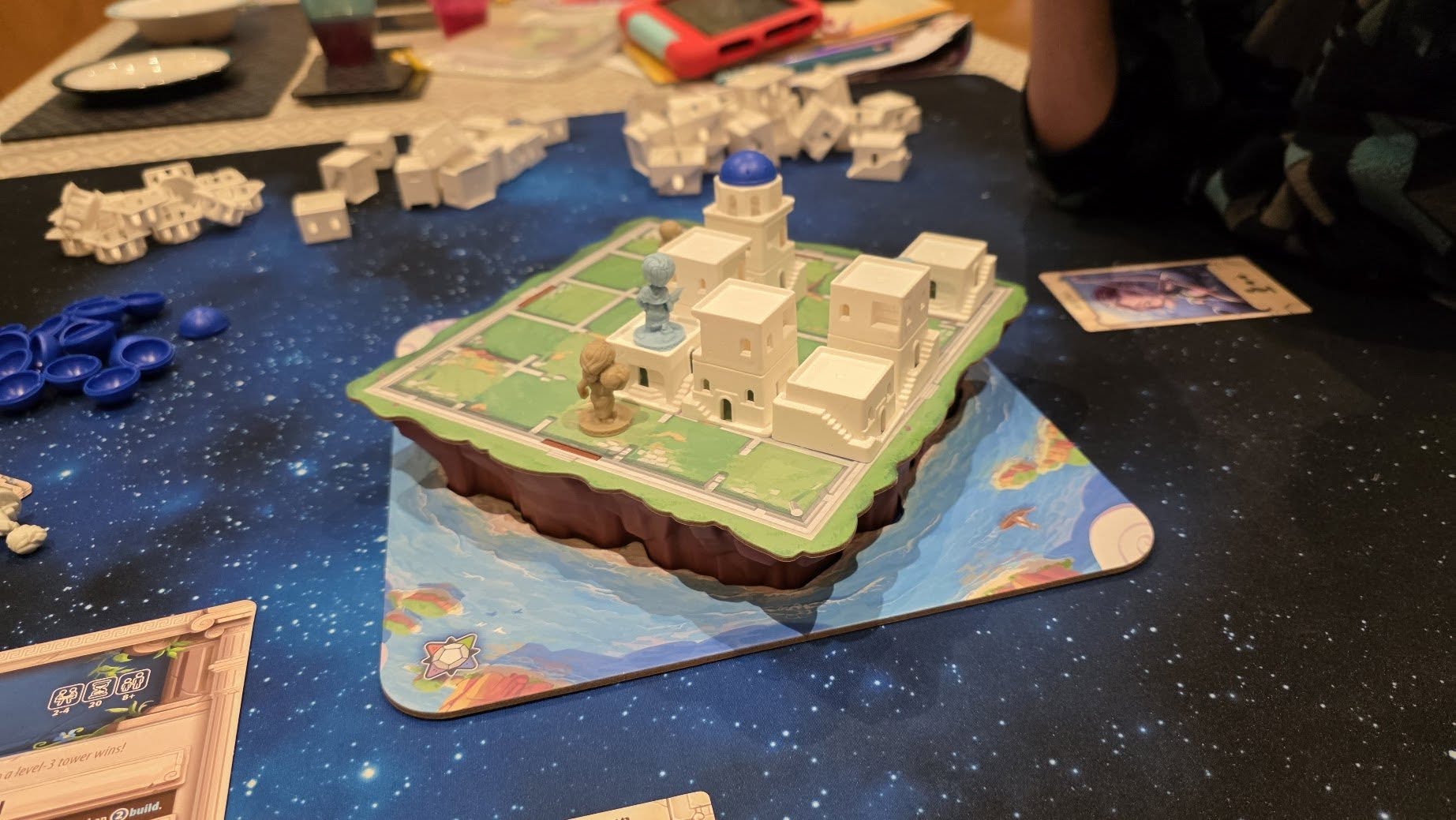 Board game setup featuring a detailed 3D model of a building layout on a cosmic-themed game mat, with various game pieces scattered around, including tokens and cards, illustrating an engaging gameplay experience.