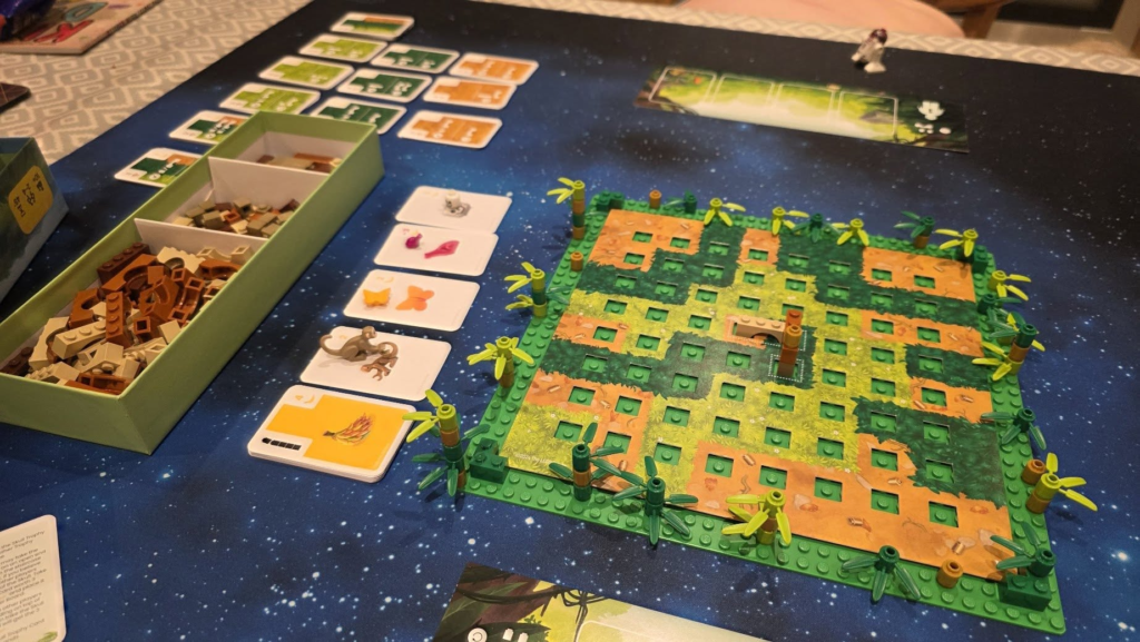 Image featuring a colorful board game setup on a galaxy-themed table. The scene includes a green game board with a grid pattern, surrounded by vibrant plastic plant pieces, alongside a box containing game pieces. Visible cards and tokens suggest gameplay elements, indicating a strategy or resource management theme.