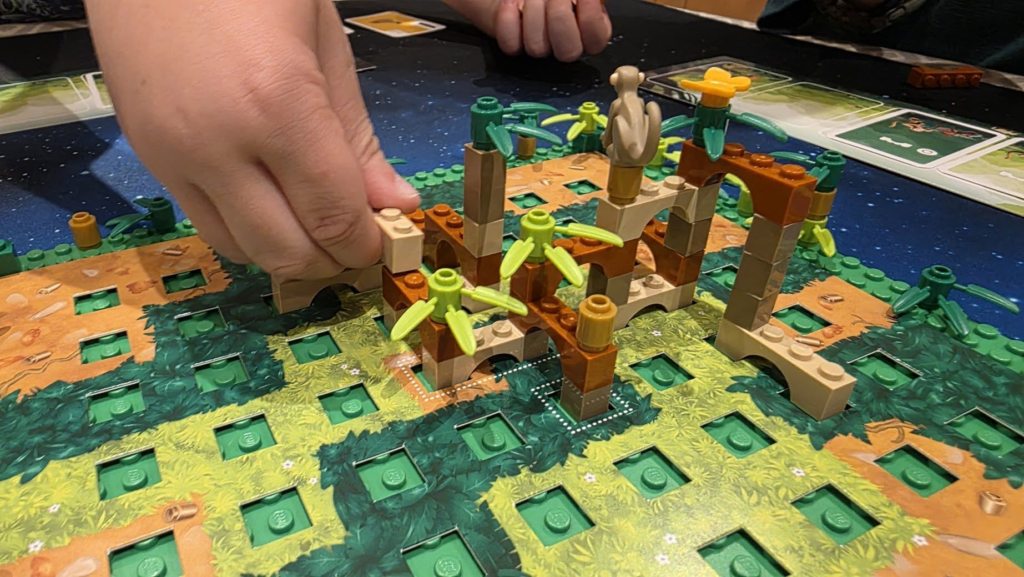 Image of a hands-on board game featuring a colorful landscape made of LEGO bricks, with a focus on a player's hand assembling a structure. The game emphasizes creativity and strategic building on a green board.